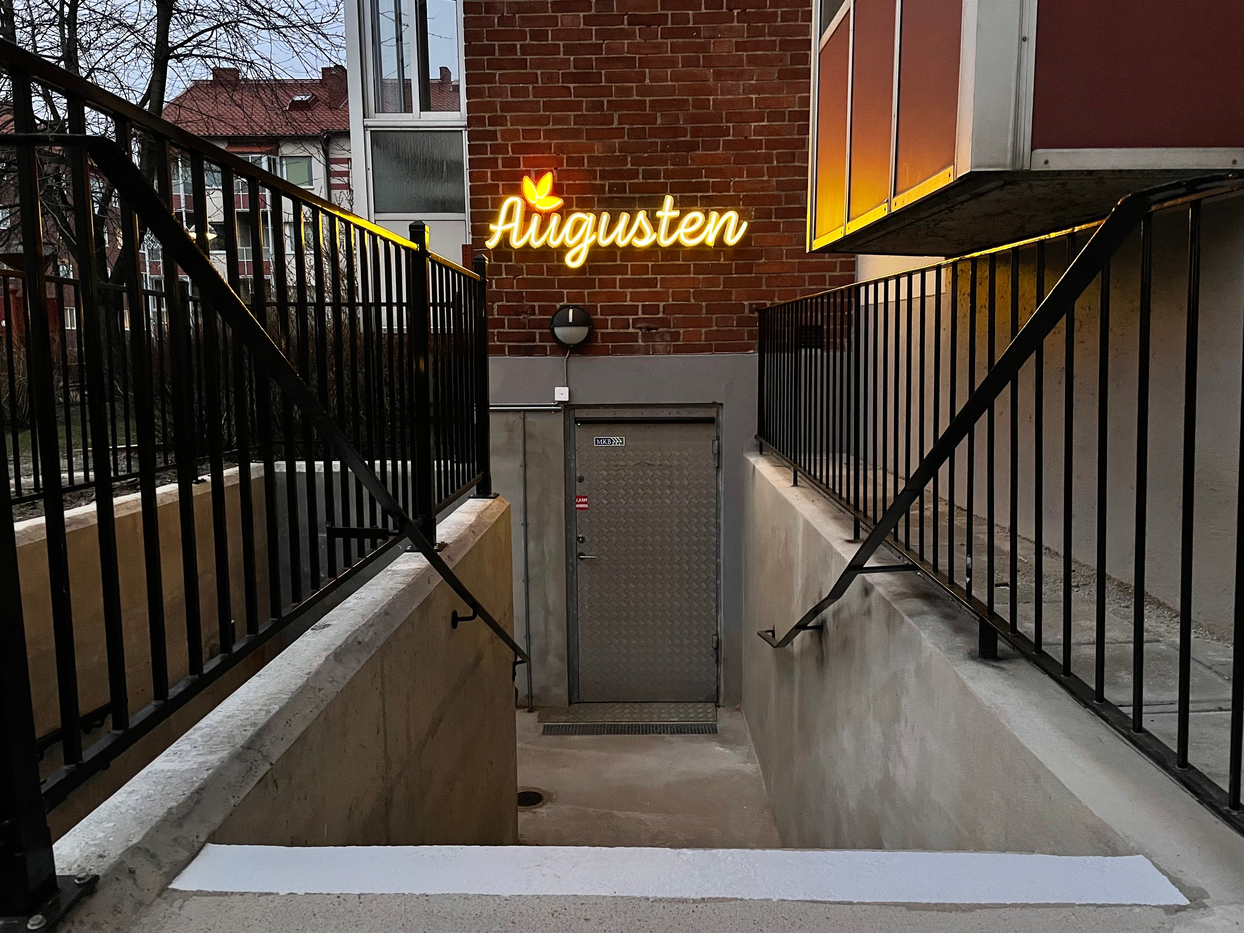 Bilden visar ingången till Augusten som är en trappa ner i en källare. Augusten skylten lyser i neon ovanför dörren.
